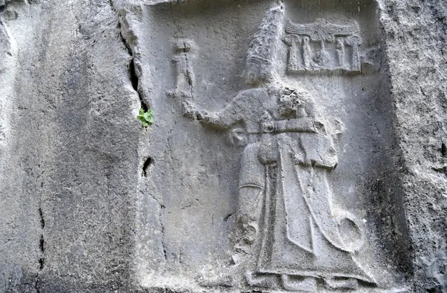 Una singola divinità ittita scolpita in un pannello rettangolare di roccia a Yazılıkaya vicino a Hattusa, Turchia, è raffigurata di profilo con un'alta corona a più corna e un bastone rituale in mano, con vesti e cintura ancora nettamente definiti nel calcare grigio.