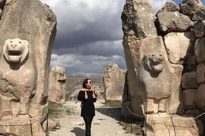 Una visitante se encuentra entre dos guardianes de piedra caliza con fauces abiertas rugiendo en la Puerta de los Leones de Hattusa, la antigua capital hitita cerca de Boğazkale, Turquía, la escala de los masivos animales de piedra enfatizando su presencia imponente sobre la antigua entrada.