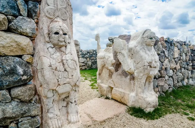 Una vista ravvicinata e angolata alla Porta del Re, Hattusa, Turchia, mostra in primo piano il pilastro guardiano con volto umano finemente scolpito, con la sfinge alata e la seconda figura guardiana visibili attraverso l'apertura della porta nell'antica cinta muraria ittita.