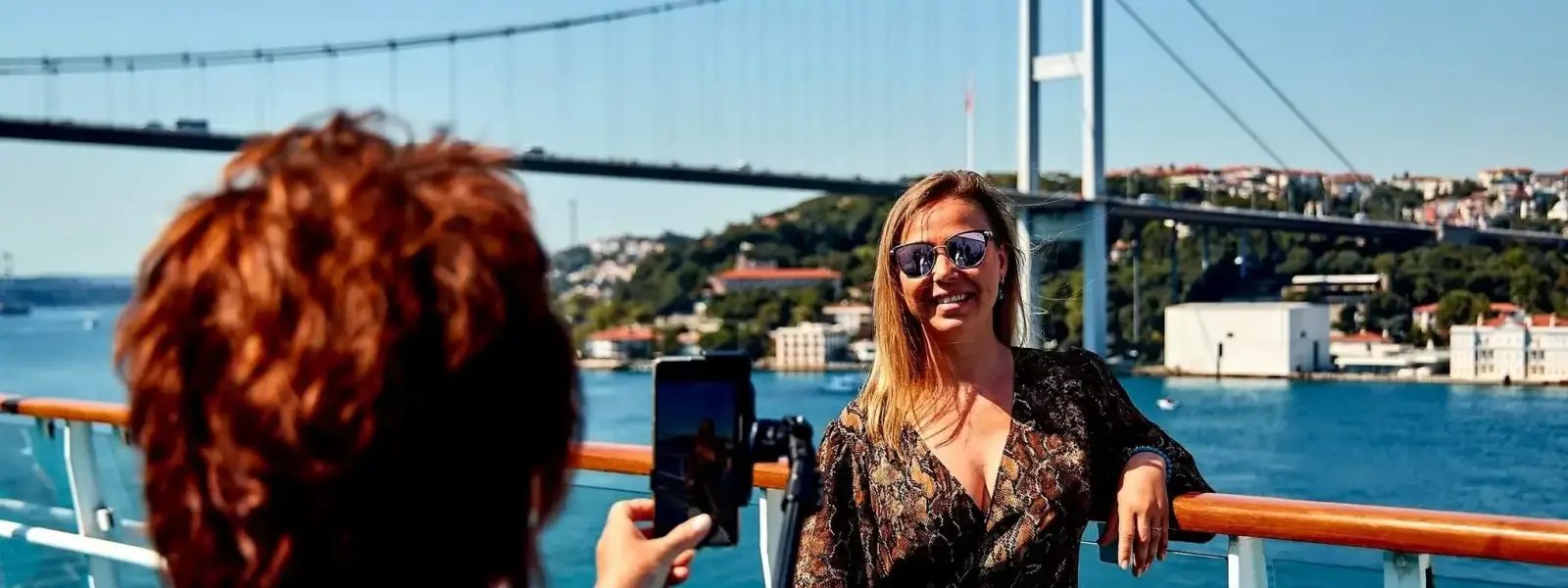 Turisti a bordo di una crociera sul Bosforo di Istanbul si riuniscono sul ponte sotto uno degli iconici ponti sospesi della città, ammirando il patrimonio culturale di questo leggendario stretto tra Europa e Asia.