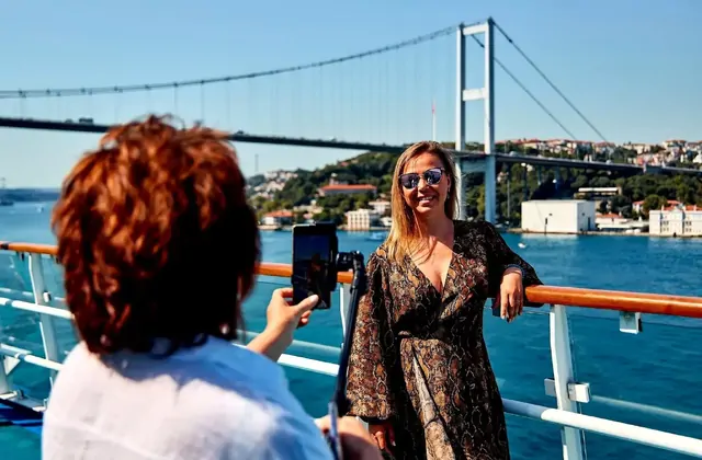 Turisti a bordo di una crociera sul Bosforo di Istanbul si riuniscono sul ponte sotto uno degli iconici ponti sospesi della città, ammirando il patrimonio culturale di questo leggendario stretto tra Europa e Asia.