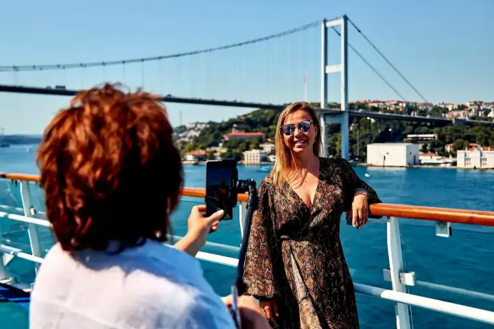 Turisti a bordo di una crociera sul Bosforo di Istanbul si riuniscono sul ponte sotto uno degli iconici ponti sospesi della città, ammirando il patrimonio culturale di questo leggendario stretto tra Europa e Asia.