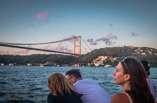 La luce dorata del tramonto illumina i passeggeri di un tour guidato in barca sul Bosforo di Istanbul mentre l'imbarcazione passa sotto un imponente ponte, tingendo lo stretto di caldi toni ambra e arancione.