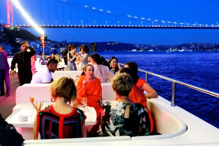Ospiti a cena sul ponte all'aperto di una crociera con cena sul Bosforo a Istanbul, circondati dai monumenti del patrimonio culturale della città illuminati lungo la riva di notte.