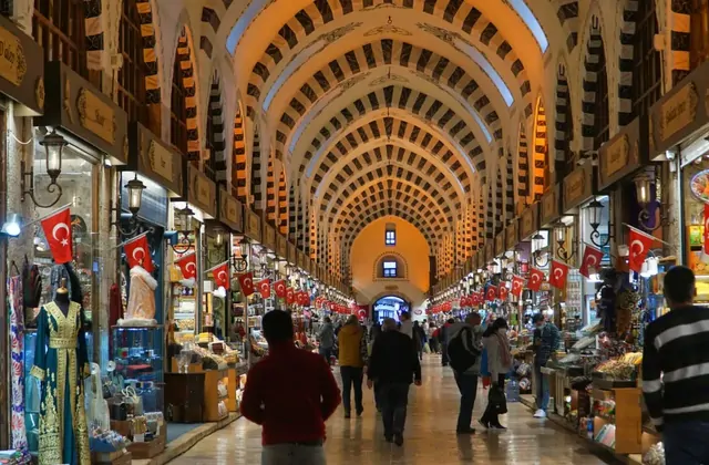 A long vaulted arcade of Istanbul's Grand Bazaar stretches into the distance, its arched brick ceiling decorated with repeating geometric patterns while Turkish flag bunting lines both sides and shoppers browse souvenir stalls in the foreground.