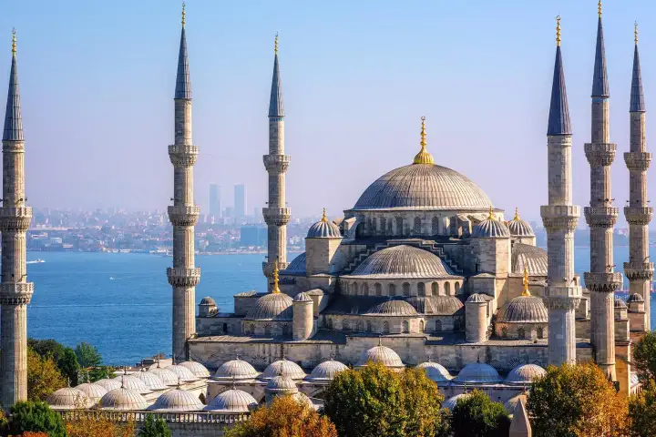 An elevated aerial view of Istanbul's Sultan Ahmed Mosque (Blue Mosque) captures all six slender minarets framing the cascading hierarchy of domes, with the blue expanse of the Bosphorus and the Asian shore of Turkey clearly visible behind the complex.