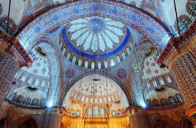 L'imponente interno della Moschea Blu (Sultanahmet) di Istanbul è ripreso guardando direttamente verso la cupola centrale, ricoperta da migliaia di piastrelle di Iznik dipinte a mano in blu e bianco con intricati motivi floreali, con semicupole e archi ogivali che si allontanano in tutte le direzion