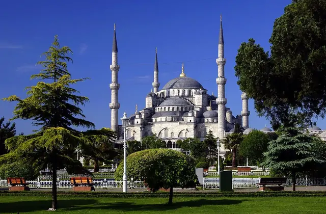 La Moschea di Sultanahmet (Moschea Blu) di Istanbul si innalza sopra un parco pubblico ben curato in una limpida giornata di sole, la sua facciata in pietra chiara e le cupole argentee che spiccano nettamente su un cielo azzurro intenso, con cipressi e cedri maturi che incorniciano il vialetto d'acc