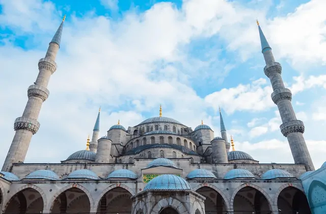 La Moschea di Sultanahmet (Moschea Blu) di Istanbul è vista dal livello del cortile guardando verso l'alto, con le cupole inferiori della sala di preghiera e gli archi del porticato che riempiono il primo piano, mentre due dei sei minareti rastremati con i loro elaborati balconi si innalzano verso u