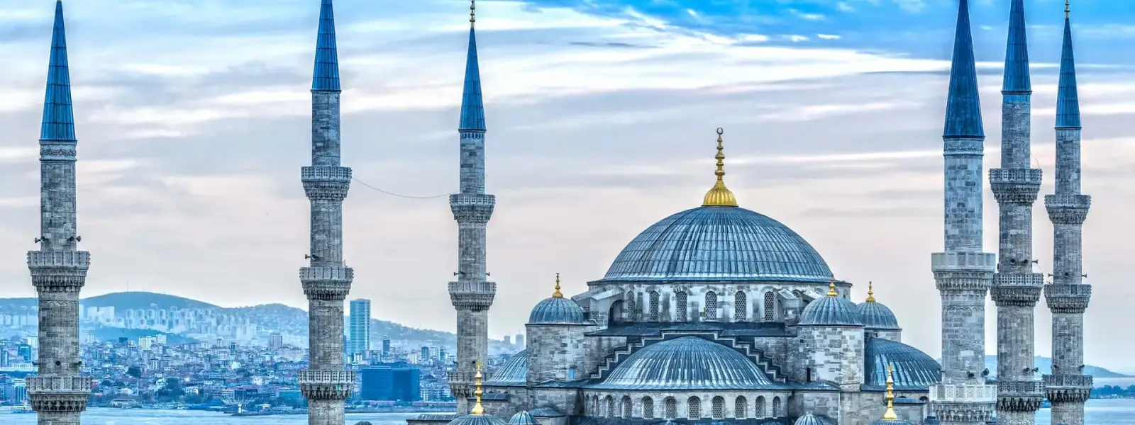 La Moschea di Sultanahmet (Moschea Blu) di Istanbul si staglia sulle case della città contro un cielo nuvoloso, con lo stretto del Bosforo visibile alle sue spalle insieme a un traghetto che attraversa l'acqua e i quartieri in collina della riva asiatica che si estendono sullo sfondo.