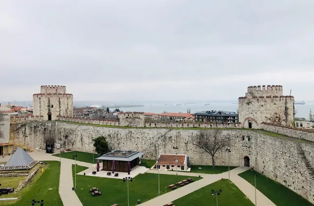 Vista a livello del suolo all'interno della storica Fortezza di Yedikule di Istanbul che mostra l'ampio cortile interno verde fiancheggiato da due massicce torri bizantine merlate, un basso edificio espositivo al centro, e oltre i merli le acque grigie del Mar di Marmara punteggiato di navi mercanti