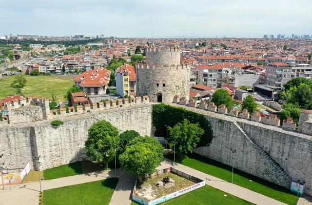 Una vista a livello drone della Fortezza di Yedikule di Istanbul (Castello delle Sette Torri) mostra un'unica alta torre cilindrica che si eleva sopra le mura merlate di collegamento, circondata da un lussureggiante giardino interno con un cartello "Yedikule Hisarı" visibile, mentre la vasta distesa