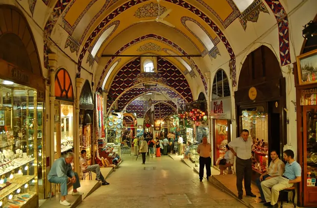 Una galleria interna del Gran Bazar di Istanbul (Kapalıçarşı) mostra un soffitto a volta ogivale riccamente dipinto con geometrie ottomane in blu e rosso, gioiellerie con vetrine in vetro illuminate lungo il lato sinistro e venditori di souvenir e tessuti a destra, con visitatori in passeggio.