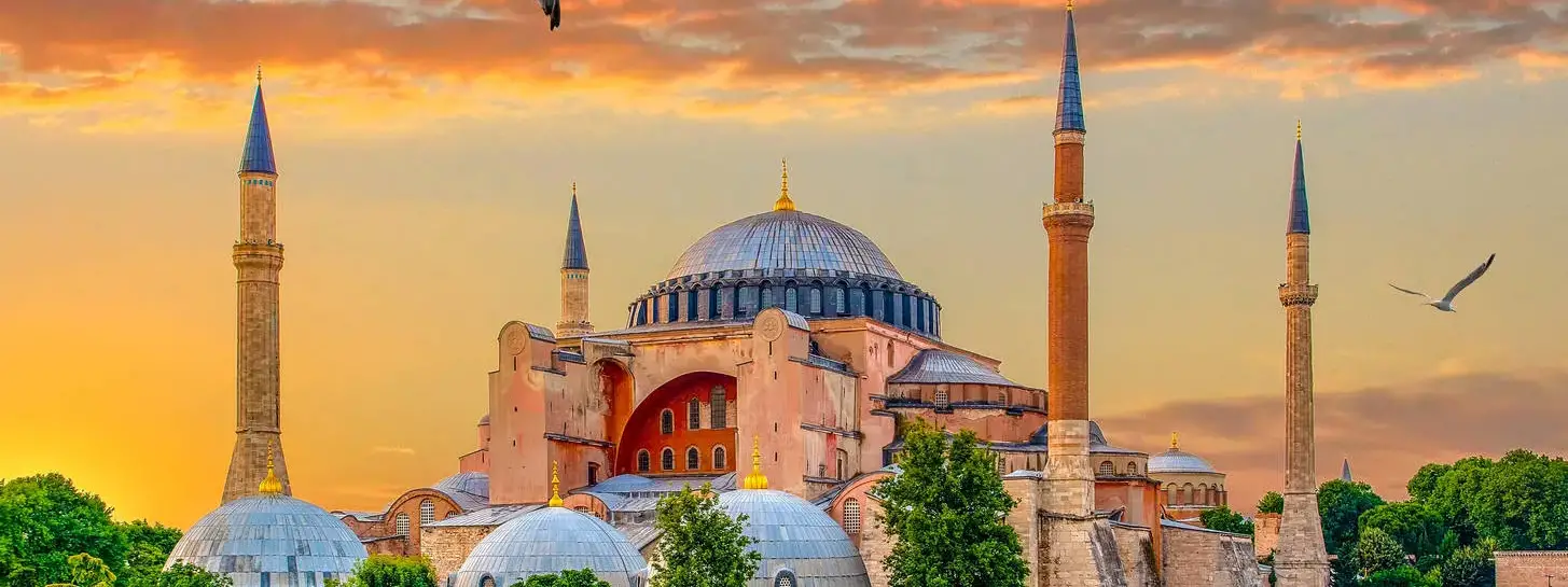 La Grande Moschea di Hagia Sophia a Istanbul splende in uno spettacolare tramonto infuocato, la sua massiccia cupola centrale e i quattro minareti ottomani che si stagliano in controluce su un cielo ardente arancione e cremisi, con i gabbiani che volano in alto e le cupole più piccole e gli alberi d