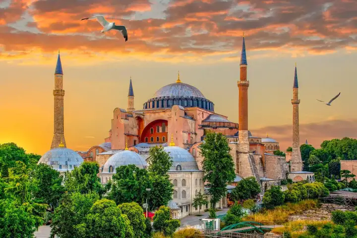 La Grande Moschea di Hagia Sophia a Istanbul splende in uno spettacolare tramonto infuocato, la sua massiccia cupola centrale e i quattro minareti ottomani che si stagliano in controluce su un cielo ardente arancione e cremisi, con i gabbiani che volano in alto e le cupole più piccole e gli alberi d
