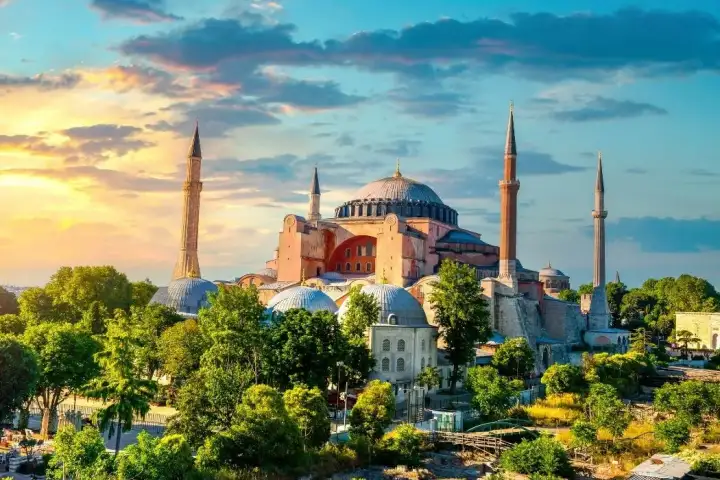 La Grande Moschea di Hagia Sophia a Istanbul è avvolta da una calda luce dorata del tramonto, le pietre marrone-rossastre delle sue antiche mura bizantine e la cupola centrale che brillano di ambra-arancio, circondata da lussureggianti alberi estivi, con un vivace cielo blu e giallo al tramonto con 