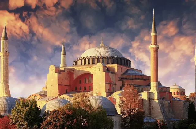 Istanbul's Hagia Sophia Grand Mosque photographed against a dramatic stormy autumn sky, its massive central dome and four pencil-thin Ottoman minarets glowing golden in the late light, the ancient red-brick Byzantine exterior walls rising from a base of smaller domes and autumn-coloured trees.