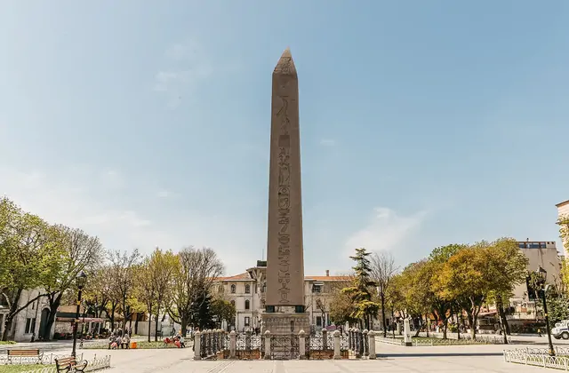 L'Obelisco di Teodosio si erge solitario in Piazza Sultanahmet a Istanbul (Ippodromo) in un limpido giorno di primavera, le sue quattro facce coperte di geroglifici egizi chiaramente leggibili mentre si affila verso la punta su un cielo azzurro pallido, con ringhiere in ferro alla base e alberi fron