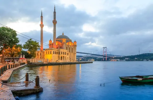 La Mezquita de Ortaköy de Estambul está iluminada con cálidas luces ámbar al anochecer, su silueta de cúpula barroco-otomana y dos esbeltos minaretes reflejándose en las oscuras aguas del Bósforo, con un pequeño barco de pesca amarrado en primer plano y el iluminado Puente del 15 de Julio arqueándos