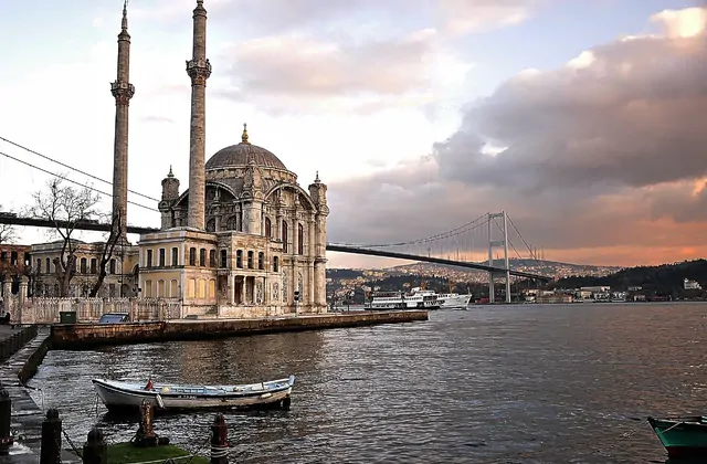 La Moschea di Ortaköy a Istanbul si erge sul lungofiume del Bosforo sotto un cielo tempestoso al tramonto, la sua sontuosa facciata in pietra e i minareti gemelli che spiccano contro le nuvole scure, con piccole barche a remi in primo piano, un traghetto passeggeri che transita al centro del canale