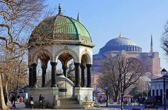 La Fontana Tedesca ottagonale (Fontana Kaiser Guglielmo II) in Piazza Sultanahmet a Istanbul è fotografata in una giornata invernale, la sua caratteristica cupola in rame verde e le arcate a colonne di marmo scuro lucidato a fuoco nitido, con la massiccia cupola di Hagia Sophia che si erge in modo p