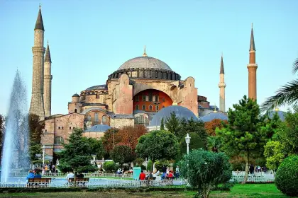 Visitors relax in the landscaped gardens in front of Istanbul's Hagia Sophia Grand Mosque on a sunny afternoon, with a decorative stone fountain and pool in the foreground, palm trees and benches lining the pathway, and the mosque's imposing Byzantine dome and Ottoman minarets rising above the summe