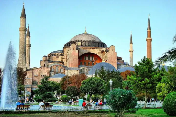 Visitors relax in the landscaped gardens in front of Istanbul's Hagia Sophia Grand Mosque on a sunny afternoon, with a decorative stone fountain and pool in the foreground, palm trees and benches lining the pathway, and the mosque's imposing Byzantine dome and Ottoman minarets rising above the summe