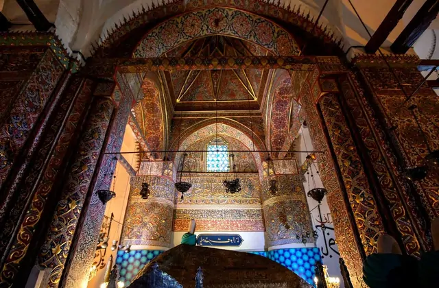 The richly gilded interior of Mevlana Museum in Konya glows with intricate mosaic tilework, carved arabesque arches, and suspended oil lamps, framing a stained-glass window deep inside the ceremonial hall.