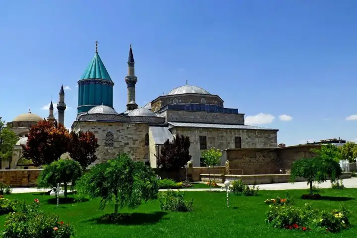 Vista panorámica del jardín del complejo del Museo Mevlana en Konya en un luminoso día de verano, con la icónica cúpula cónica turquesa elevándose detrás del edificio de la mezquita de piedra y dos esbeltos minaretes apuntando al cielo azul intenso.