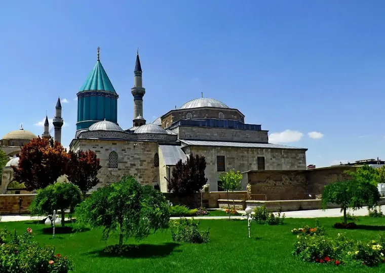 A wide-angle garden view of the Mevlana Museum complex in Konya on a bright summer day, with the iconic turquoise conical dome rising behind the stone mosque building and two slender minarets pointing into the deep blue sky.