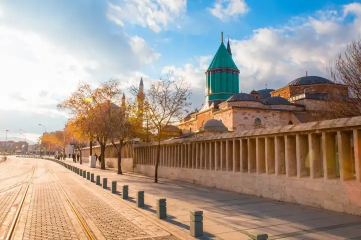 The long colonnaded outer wall of the Mevlana Museum complex stretches along a wide cobblestone boulevard in Konya at golden hour, with the turquoise fluted dome and mosque visible above the roofline against a partly cloudy autumn sky.