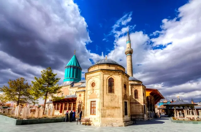 El patio exterior del complejo del Museo Mevlana en Konya bajo un cielo HDR vibrante, con la mezquita octagonal de ventanas arqueadas, un esbelto minarete y la brillante cúpula cónica verde azulado de la tumba de Rumi asomando sobre la línea de tejados, mientras un pequeño grupo de visitantes se enc