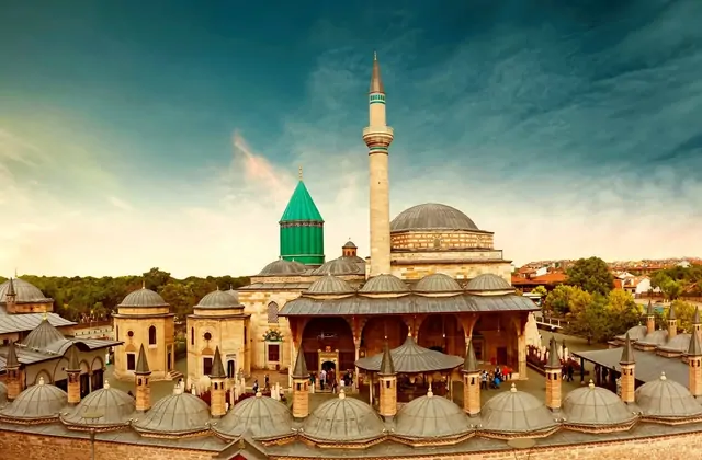 A warm sunset panorama of the Mevlana Museum complex in Konya taken from an elevated vantage point, revealing the full sweep of lead-domed portico bays, the central fountain courtyard bustling with visitors, the main mosque with its large dome and soaring minaret, and Rumi's green conical tomb glowi