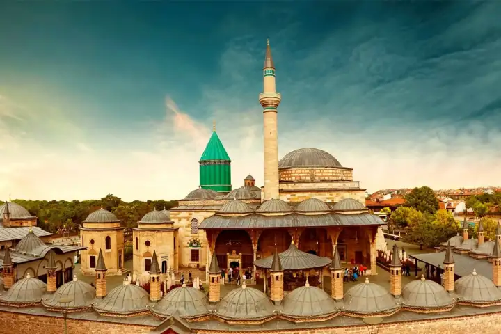 Un caldo panorama al tramonto del complesso del Museo Mevlana a Konya ripreso da un punto sopraelevato, che rivela l'intera distesa dei portici con cupole di piombo, il cortile centrale della fontana affollato di visitatori, la moschea principale con la sua grande cupola e il minareto svettante, e l