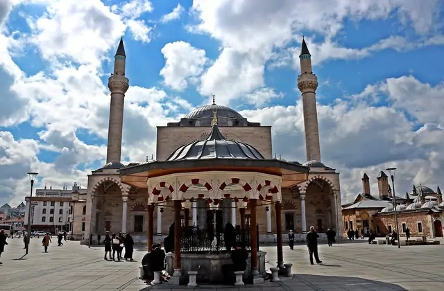 Los visitantes deambulan por la amplia plaza pavimentada frente a la Mezquita Selimiye de Konya, cuyos minaretes gemelos y cúpula central dominan el encuadre, con una gran fuente de ablución ornamentada bajo un dosel en primer plano y las cúpulas más pequeñas del adyacente complejo del Museo Mevlana