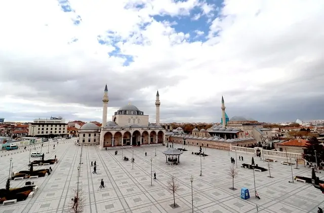 Vista panorámica elevada de la Plaza Central Alaaddin de Konya, mostrando la Mezquita Selimiye con su arcada de triple cúpula y minaretes gemelos a la izquierda, el pequeño pabellón octagonal de la fuente al centro, y el complejo del Museo Mevlana con sus tres minaretes y la lejana cúpula cónica ver
