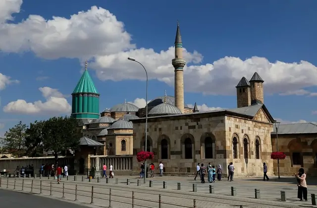 Vista a nivel de calle de la fachada frontal del complejo del Museo Mevlana en Konya, con peatones cruzando la plaza, la cúpula cónica verde turquesa de la tumba de Rumi destacada a la izquierda, la mezquita adyacente y su alto minarete al centro, y cestas de flores rojas colgando a lo largo del mur