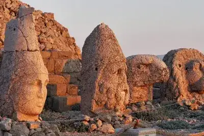 Four colossal stone heads — including a finely carved deity face with a tiara crown and the weathered head of a lion — rest on Mount Nemrut's terrace bathed in soft warm sunset light, the ancient Commagene sanctuary glowing in shades of amber across Adıyaman, Turkey.