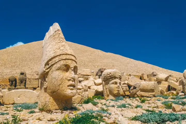 A towering stone head wearing a pointed tiara dominates the foreground of Mount Nemrut's west terrace against a brilliant blue sky, with the massive gravel tumulus of King Antiochus I rising behind and several other fallen heads scattered across the rocky slope in Adıyaman, Turkey.
