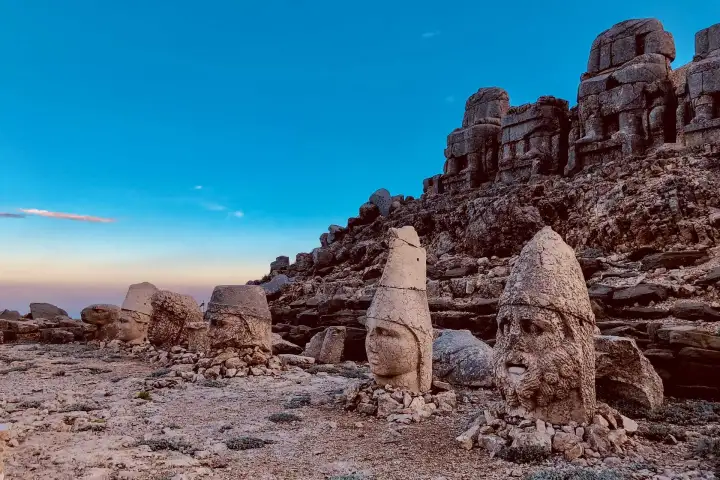 Taken at pre-dawn twilight, two freestanding stone heads — one helmeted deity and one bearded figure — stand on Mount Nemrut's east terrace as a deep cobalt sky fades to orange on the horizon, the seated headless thrones still visible on the ridge above Adıyaman, Turkey.