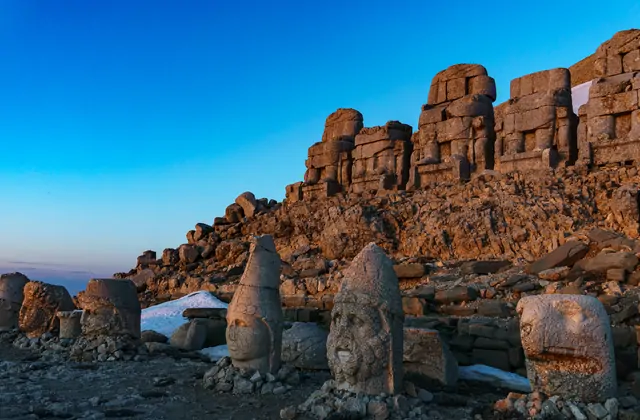 Under an electric-blue twilight sky, a row of severed stone heads on Mount Nemrut's east terrace — including a deity with a tiara and a bearded figure — are lit by warm artificial light, their conical crowns silhouetted against the deep blue horizon as the seated statues loom on the ridge above in A