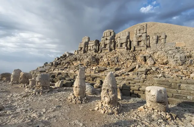 A broad, ground-level perspective of Mount Nemrut's east terrace captures three detached stone heads in the foreground — their expressions worn smooth by centuries of wind — while the full row of seated Commagene deity statues remains intact on the platform above, dramatic storm clouds gathering ove