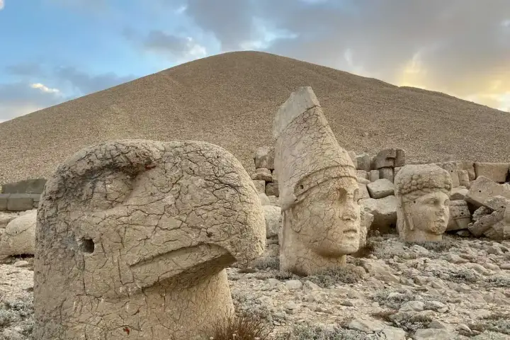 Close-up view of the eagle head sculpture and a crowned deity head on Mount Nemrut's west terrace at dusk, the eagle's beak and eye deeply carved in weathered limestone, the conical tiara of the deity sharp against a pastel sky at sunset, with the stone tumulus of Antiochus I rising behind in Adıyam