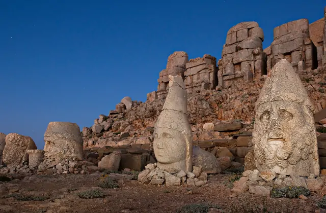 Five severed stone heads line the foreground of Mount Nemrut's east terrace in deep evening blue light, the two nearest showing a pointed-tiara deity and a bearded god in crisp detail, while the row of intact throne statues towers above them on the ridge in Adıyaman, Turkey, a single faint star visi