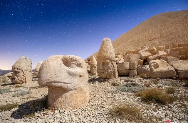Under a moonlit starry sky on Mount Nemrut, the large stone eagle head dominates the foreground with its beak open and eye socket deeply carved, while behind it the crown-topped heads of Antiochus I and other Commagene deities recede toward the gravel tumulus, the Milky Way visible over Adıyaman, Tu