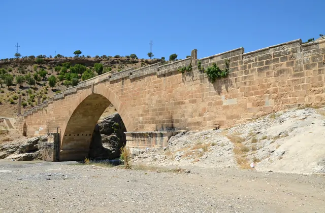 A single-arch ancient Roman bridge spans a dry riverbed in southeastern Turkey on the road to Nemrut Dağı, its honey-colored stone parapet glowing in summer heat, an ancient column standing at the near end and scrubby green vegetation framing the limestone canyon walls of the Commagene region, Adıya