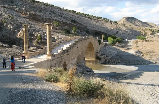 The Cendere Bridge near Nemrut Dağı photographed from river level shows its full single Roman arch with two tall Corinthian columns flanking the entrance, the warm limestone structure reflected in the slow-moving river beneath overcast autumn skies in the Commagene region of Adıyaman, Turkey.