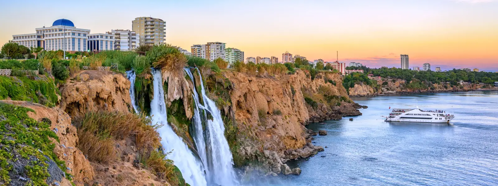 La cascada inferior de Düden en Antalya al atardecer, con cálidos tonos naranjas y rosas tiñendo el cielo sobre la ciudad, un gran crucero blanco navegando por el mar tranquilo abajo y un edificio cívico con cúpula iluminado en lo alto del acantilado.
