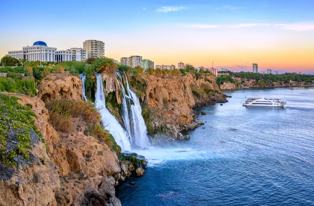 La cascata inferiore del Düden ad Antalya al crepuscolo, con caldi toni arancioni e rosa che dipingono il cielo sulla città, una grande nave da crociera bianca che naviga nel mare calmo sottostante e un prominente edificio civico con cupola illuminato in cima alla scogliera.