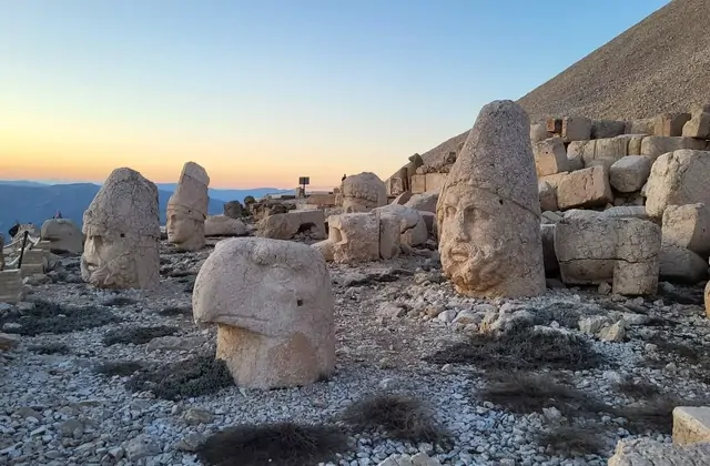 At sunrise on Mount Nemrut's west terrace, a wide scatter of severed stone heads — including a tiara-crowned deity and an eagle head — rests across the gravel plateau, the warm peach-and-orange glow of dawn low on the distant horizon and the stone tumulus of King Antiochus I silhouetted to the right